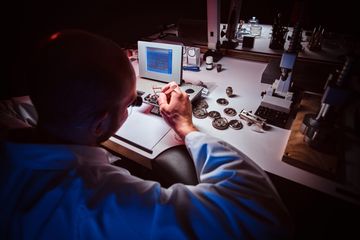 Watchmaker viewed from behind while working at a microscope and timing bench