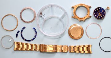 Disassembled gold Rolex Submariner with movement, bracelet, case, dial, and bezel parts on a workshop mat